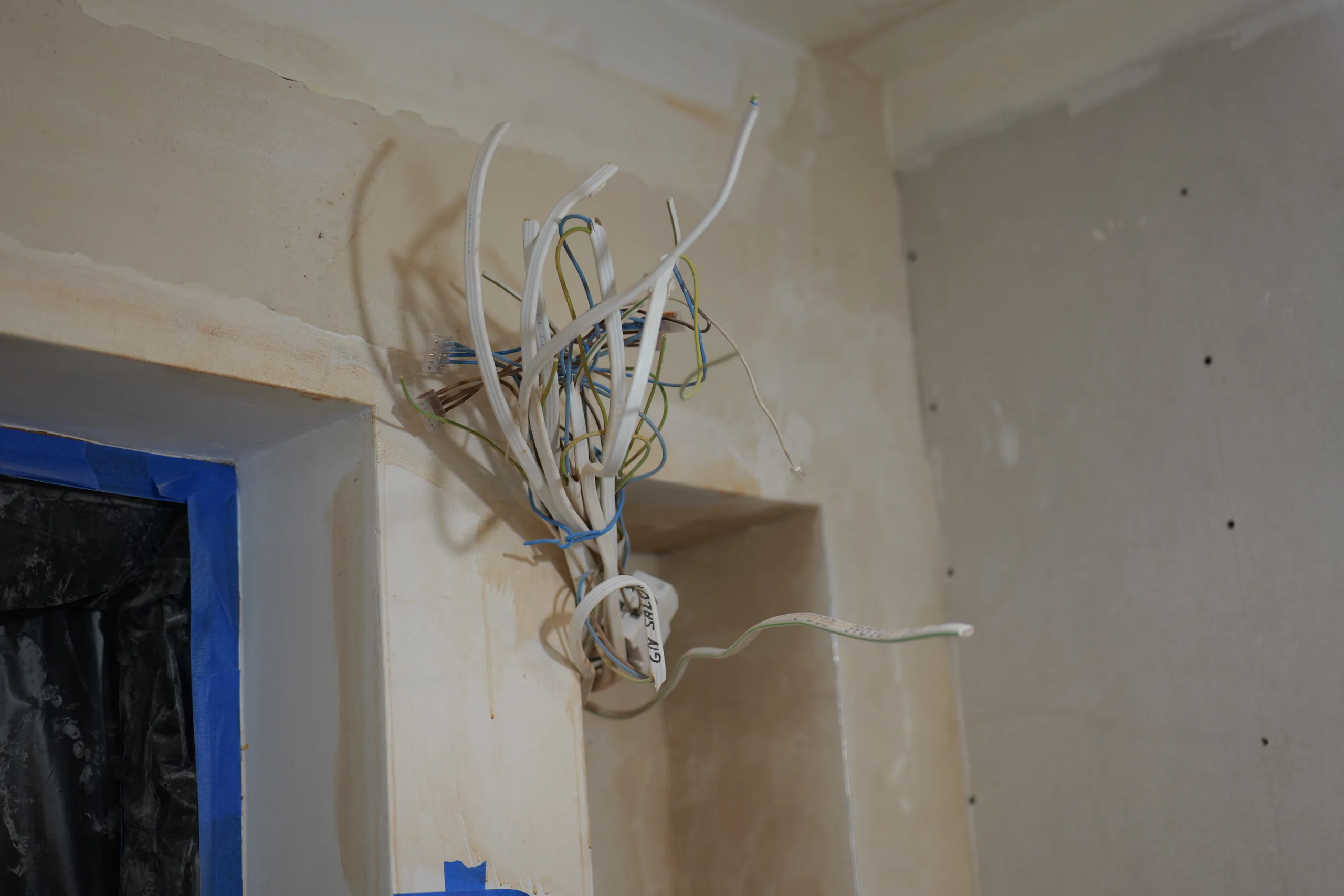 Wiązka nowych przewodów elektrycznych zwisająca z narożnika sufitu w surowym pomieszczeniu, część żył połączona kostkami, jeden przewód oznaczony ręcznie opisaną taśmą, ślady po skutym tynku na ścianach dookoła