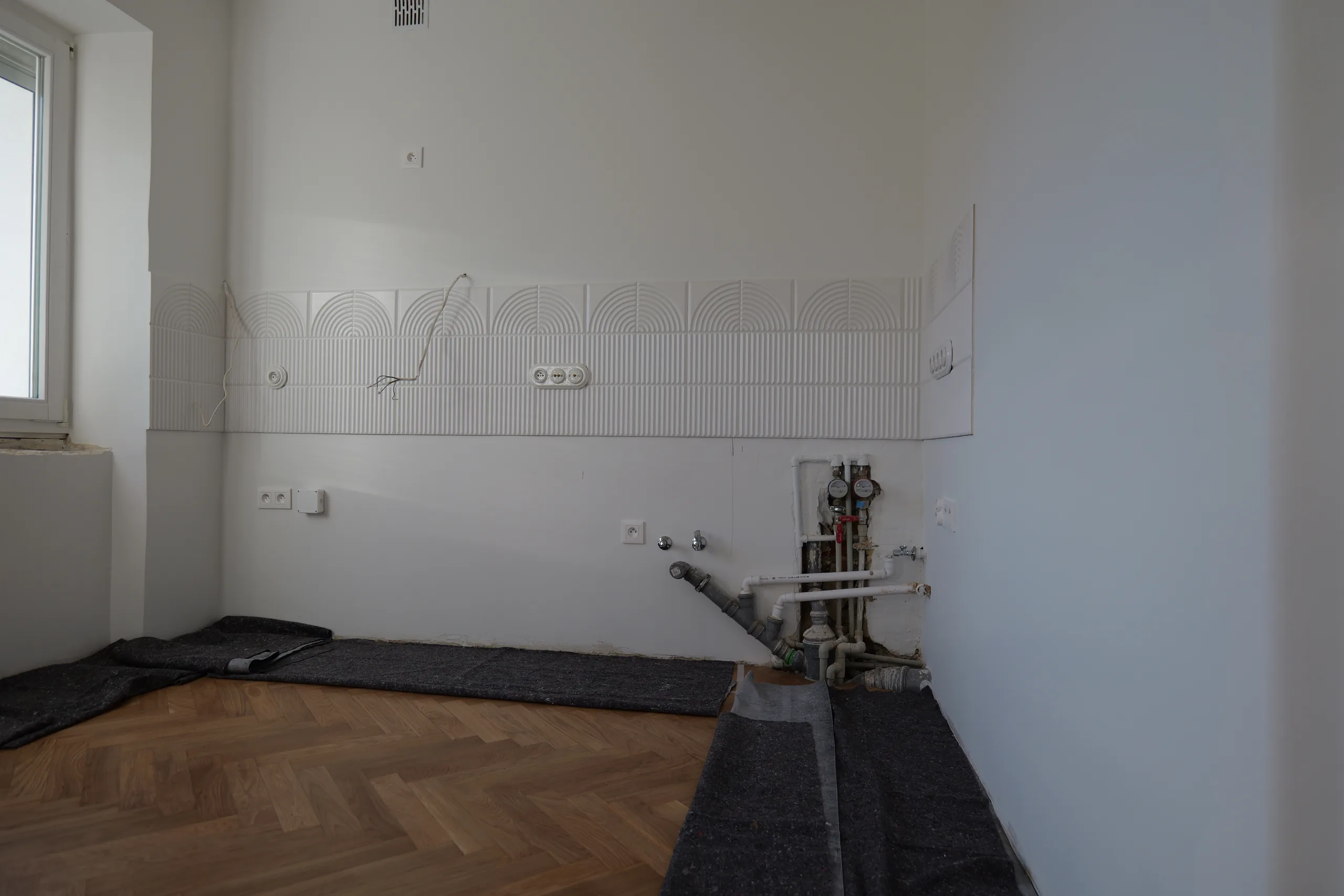 Kuchnia przed montażem zabudowy: biały dekoracyjny fartuch 3D we wzór łuków w poziomym pasie na ścianie, pod nim odsłonięte przyłącza wodne z rozdzielaczem i manometrami, gniazda elektryczne, parkiet w jodełkę przykryty matami ochronnymi