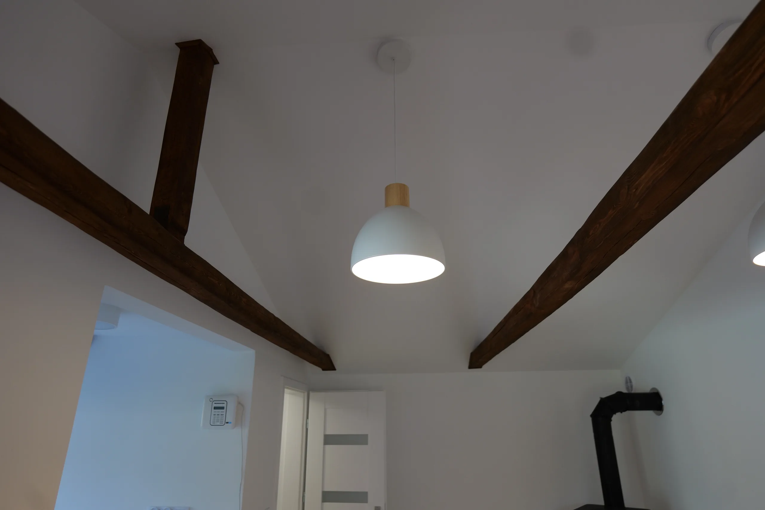 Wnętrze pod skosem dachu: ciemne bejcowane belki więźby na białym suficie, dwie białe lampy wiszące z drewnianym uchwytem, piec na drewno z czarną rurą wyprowadzoną przez sufit, podłoga w kolorze drewna